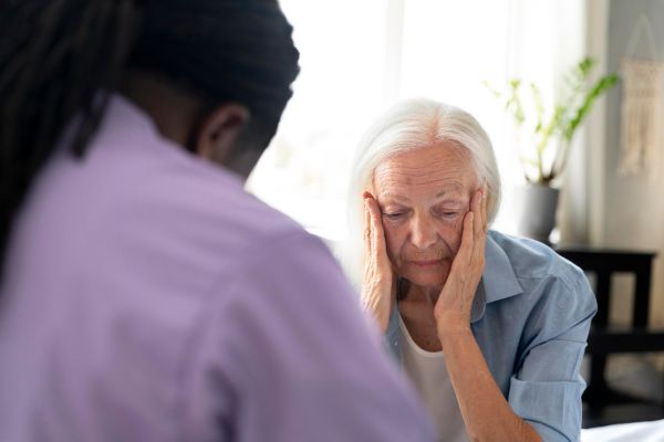 Femme âgée, tête baissée et mains sur les joues, face à une personne vue de dos